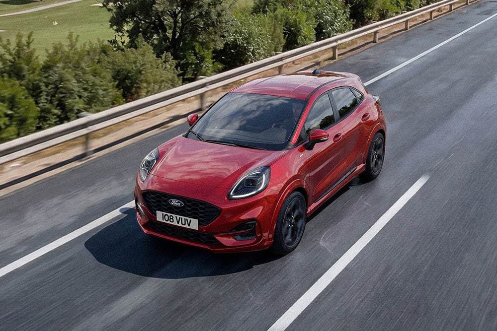 Un Ford Puma rouge roulant sur une route au milieu d'un paysage verdoyant.