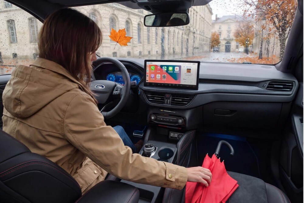 Une photo de l'intérieur d'un véhicule Ford en automne. Une femme vêtue d'une veste marronne est assise au volant et tend la main vers un parapluie rouge posé sur le siège passager.