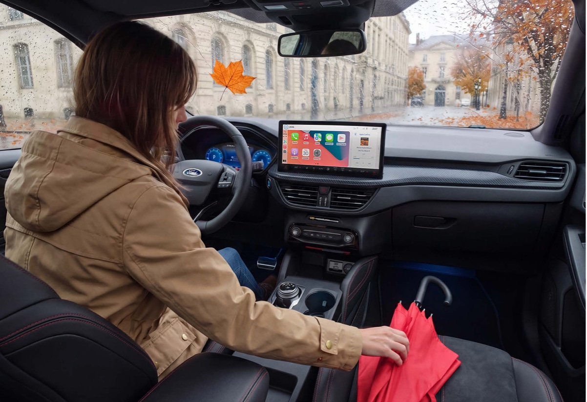 Une photo de l'intérieur d'un véhicule Ford en automne. Une femme vêtue d'une veste marronne est assise au volant et tend la main vers un parapluie rouge posé sur le siège passager.