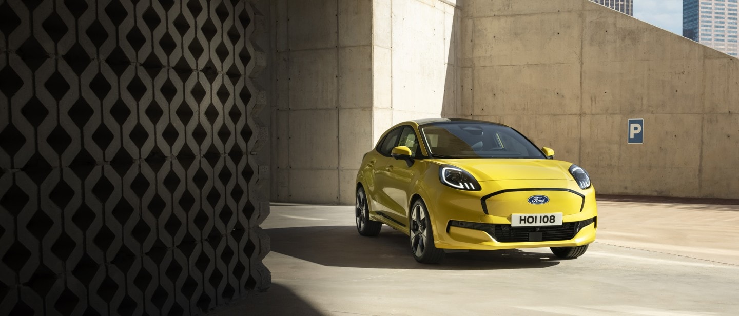 Une Ford Puma Gen-E jaune vif est garé sur une surface en béton sous une architecture moderne et angulaire en béton.