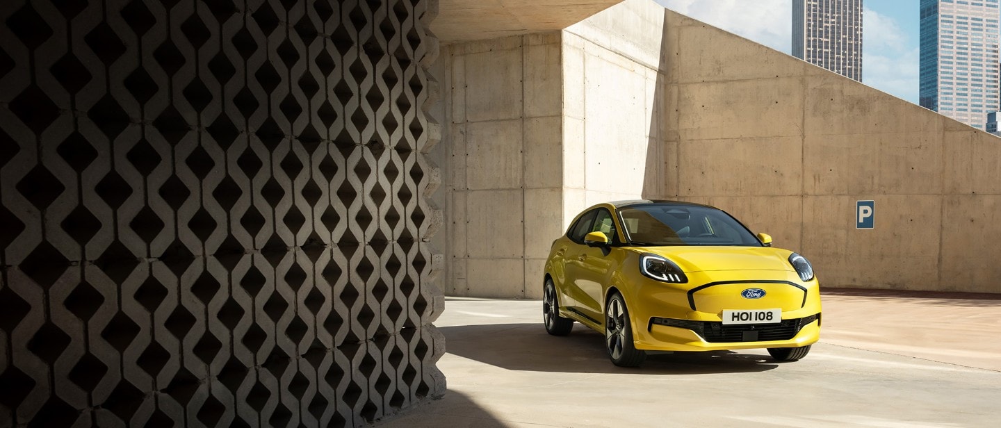 Une Ford Puma Gen-E jaune vif est garé sur une surface en béton sous une architecture moderne et angulaire en béton.