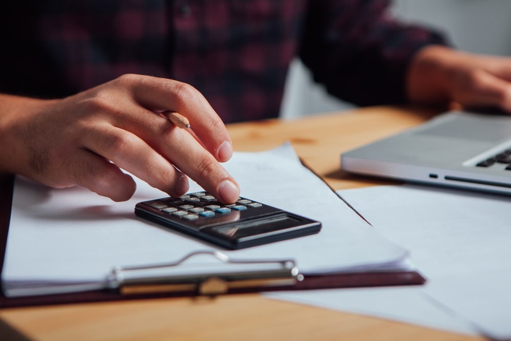Une main tape sur une calculatrice posée sur des documents, un ordinateur flou à l'arrière-plan.