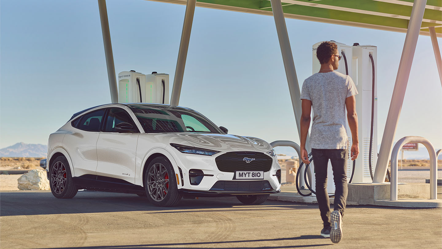 Een witte Ford Mustang Mach-E geparkeerd bij een openbaar laadstation in de buitenlucht. Een man in een wit T-shirt loopt naar de auto.
