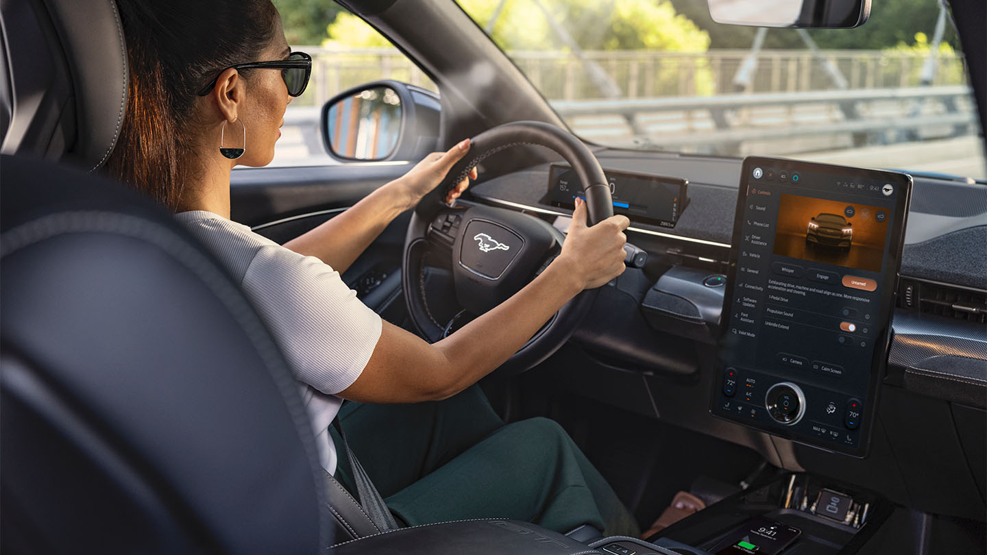 Een vrouw bestuurt de Mustang Mach-E met haar handen aan het stuur. Het 15.5" touchscreen toont details over de auto.