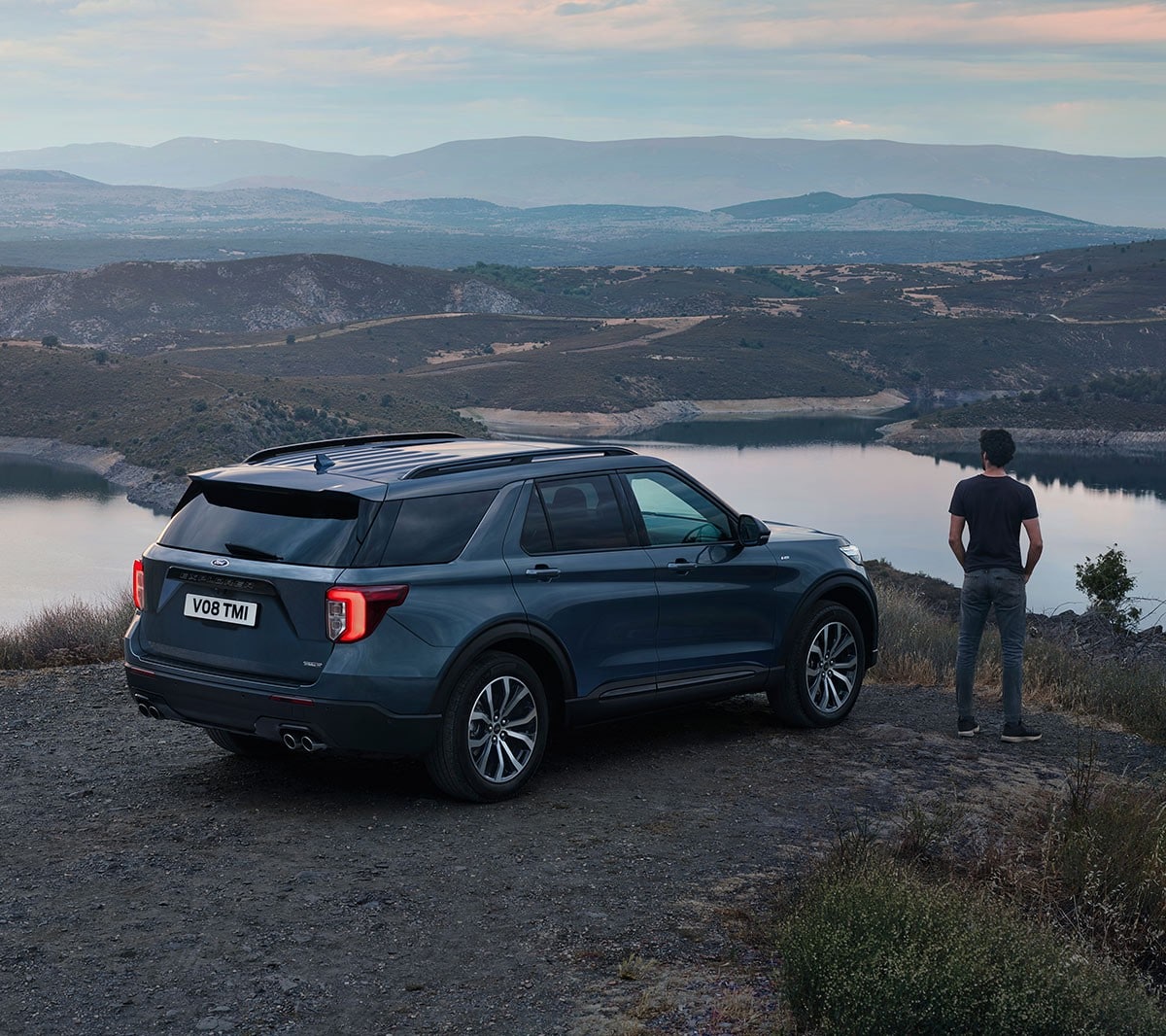 Ford Explorer parked in hilly area above lakes with person standing next to it overlooking landscape