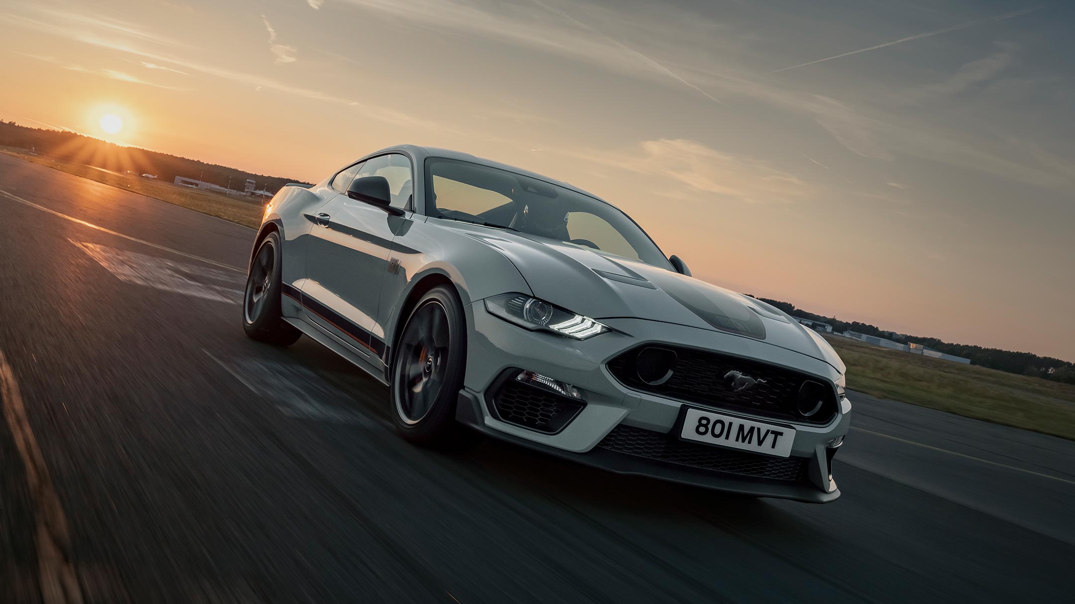 Ford Mustang Mach 1 drving on a racetrack against a sunset backdrop.