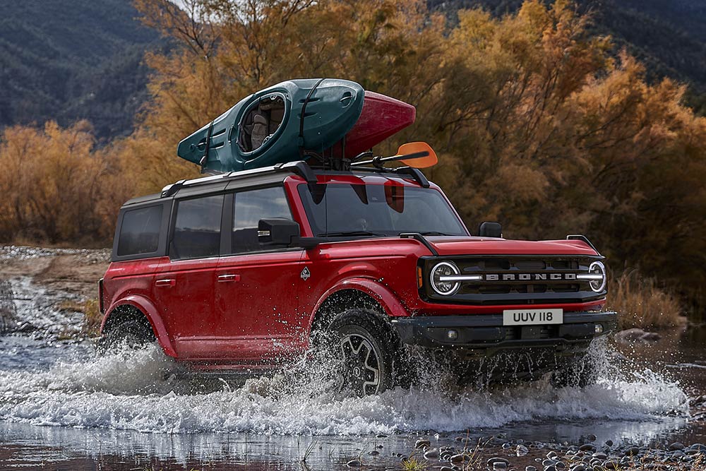 Vue latérale d’un Ford Bronco rouge traversant des eaux peu profondes en montagne.