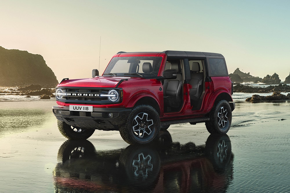 Vue latérale d’un Ford Bronco rouge, sans portes, stationné sur une rive de sable humide.