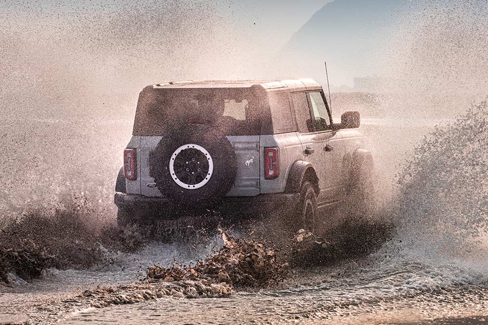 Vue arrière d’un Ford Bronco argenté traversant des embruns sur une plage.