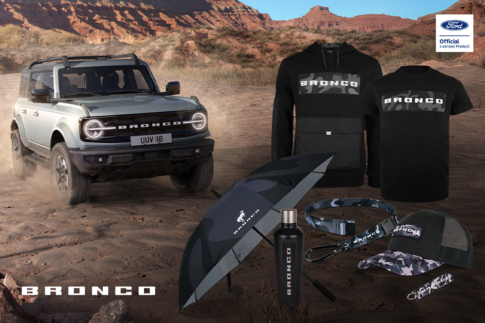 Collection Ford Bronco avec t-shirt noir, veste, polo, casquette, bonnet, sac étanche et un Bronco gris en arrière-plan.