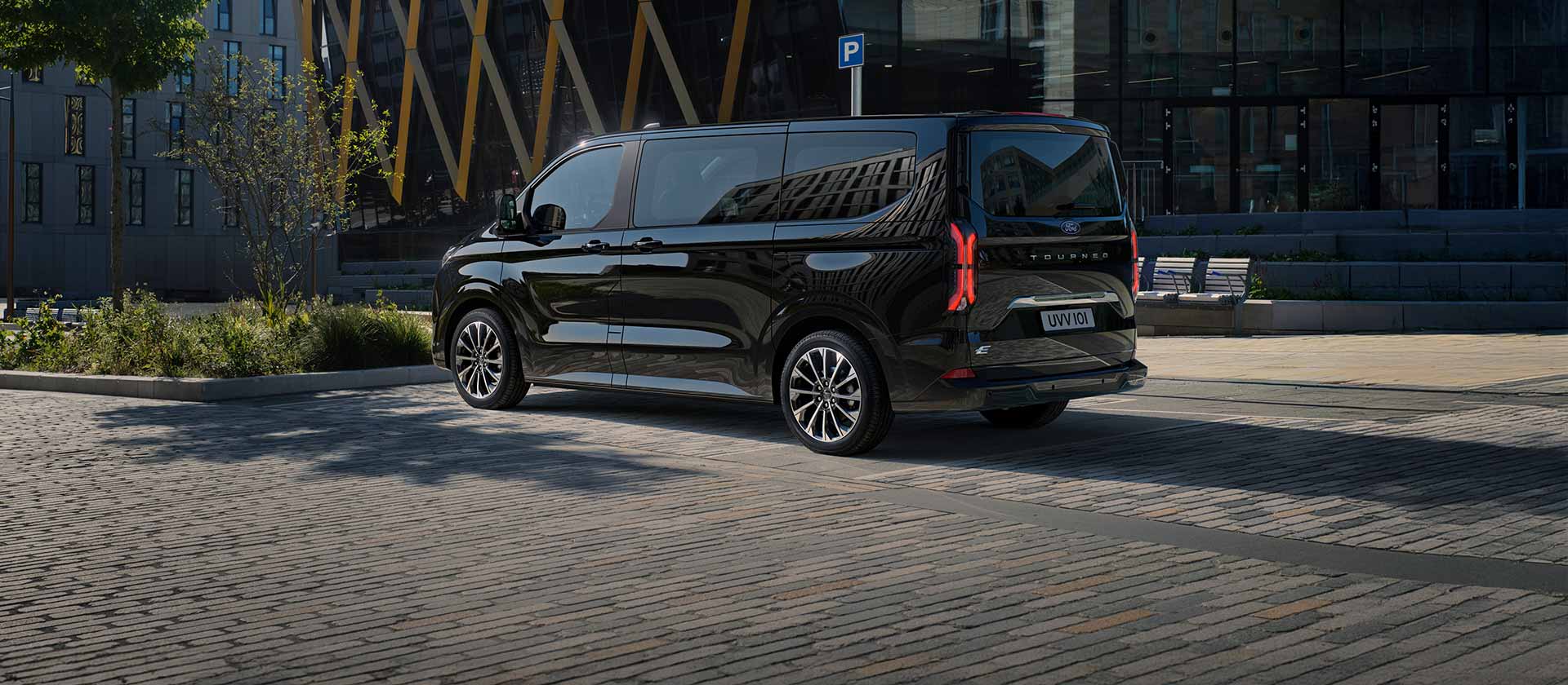 A black Ford E-Tourneo Custom is parked showing the side and rear in front of a modern building.