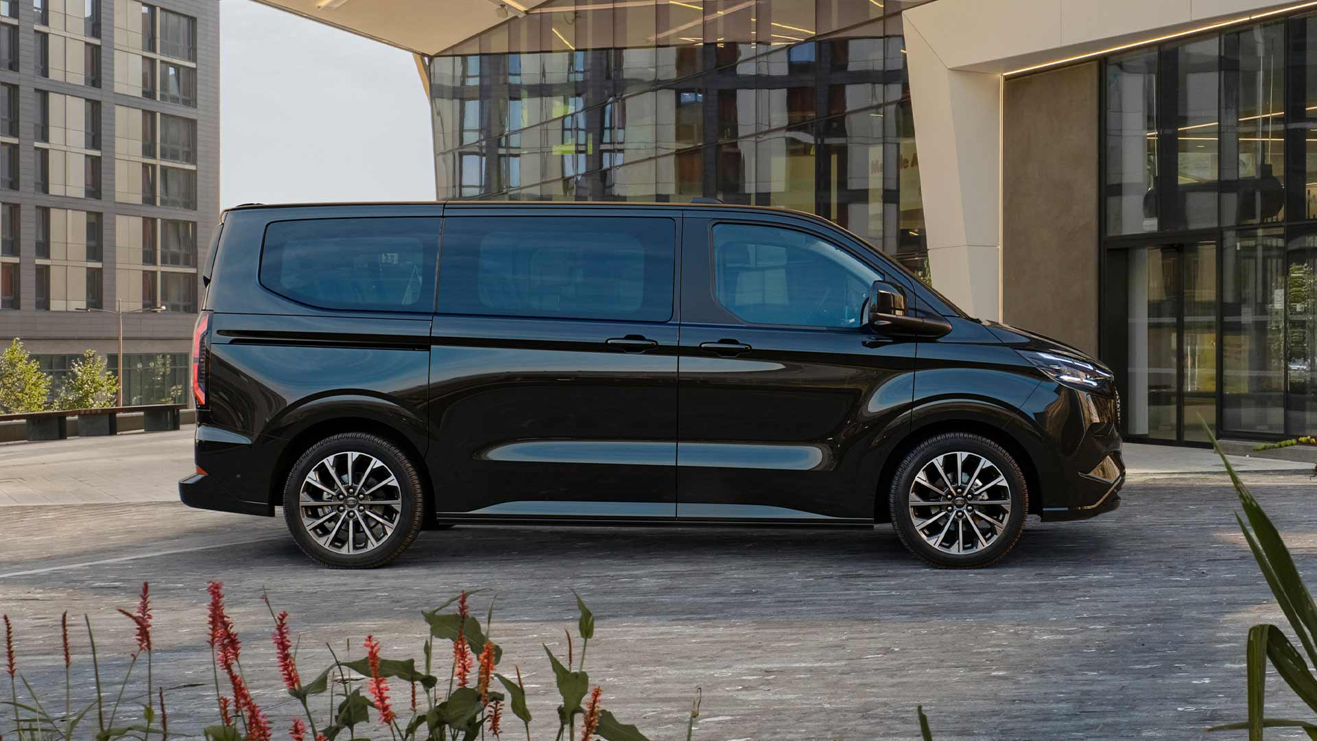Un Ford E-Tourneo Custom noir est stationné devant un bâtiment moderne.