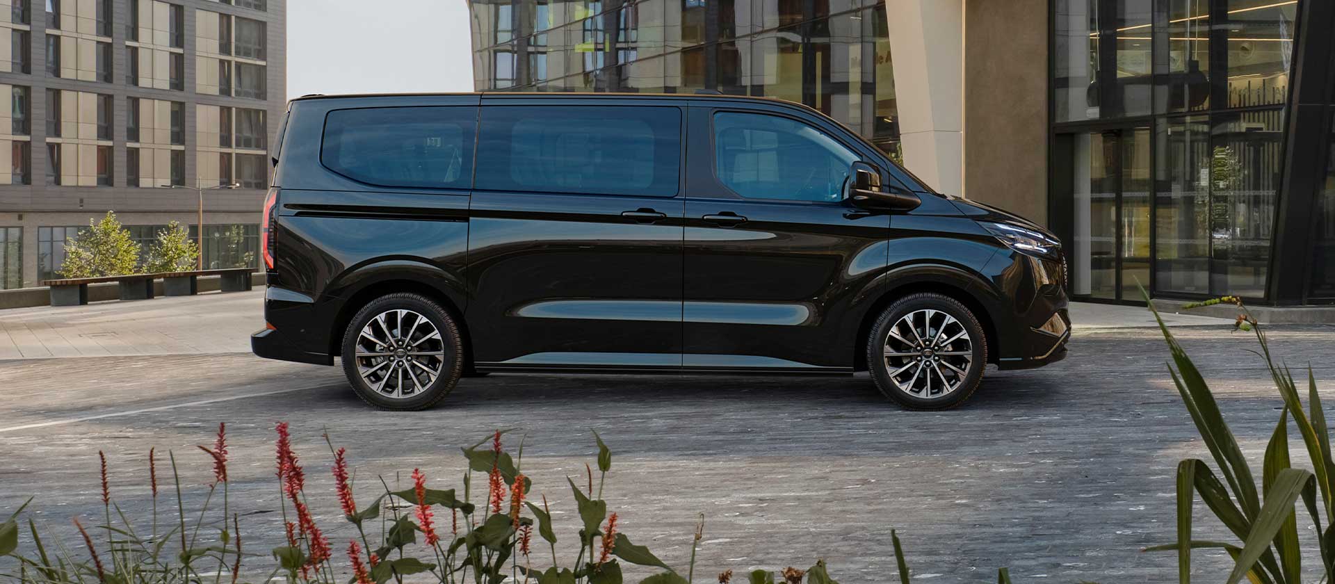 Un Ford E-Tourneo Custom noir est stationné devant un bâtiment moderne.