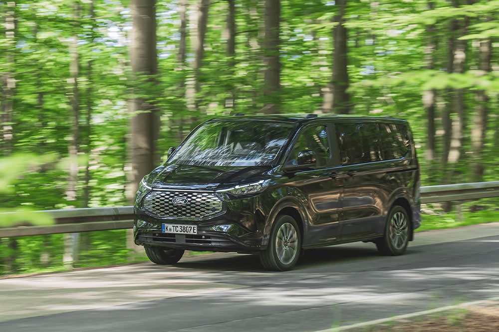 Un Ford E-Tourneo Custom noir circule sur une route bordée d’arbres.