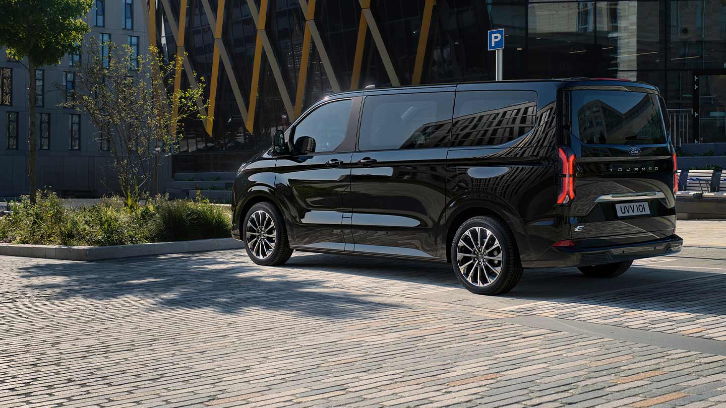 Un Ford E-Tourneo Custom noir est stationné, montrant le côté et l’arrière devant un bâtiment moderne.