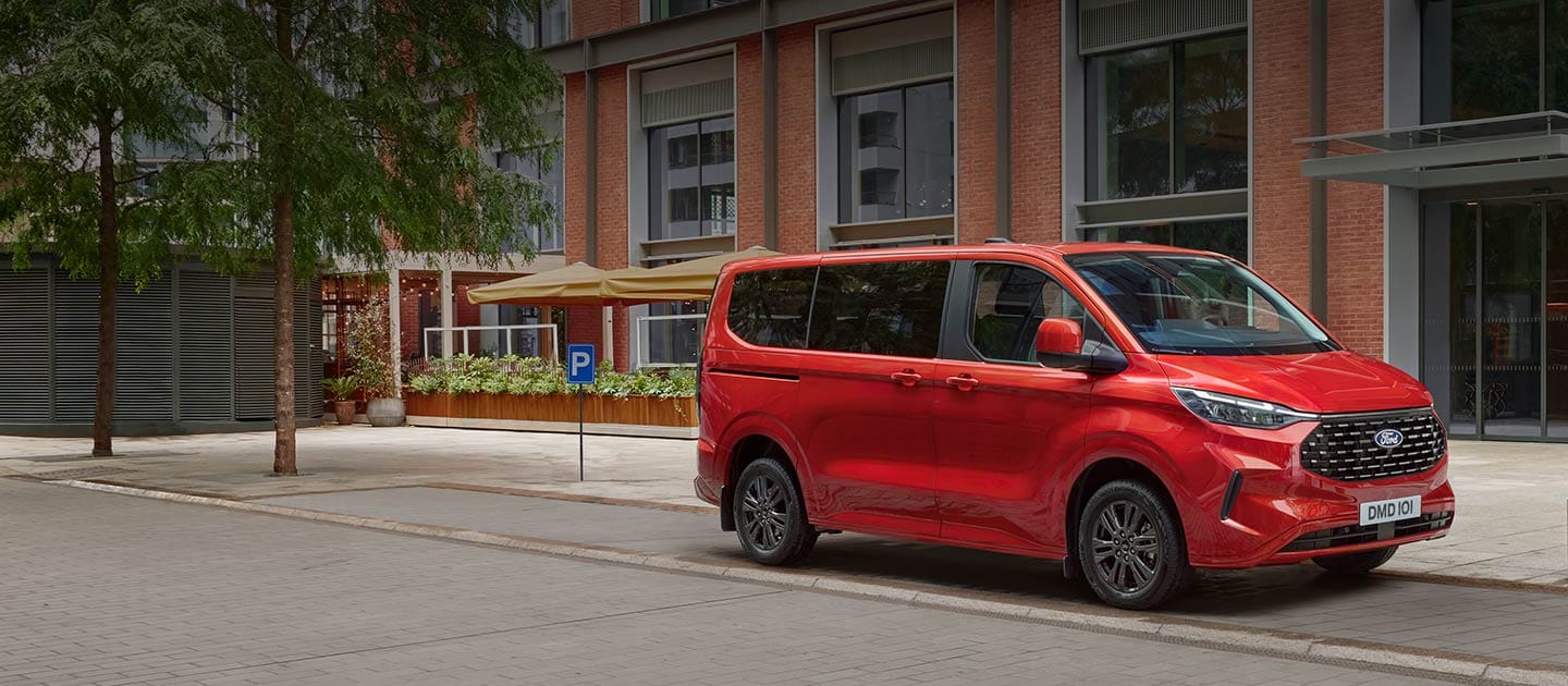 Un Ford Tourneo Custom rouge est stationné dans la rue d’une ville.