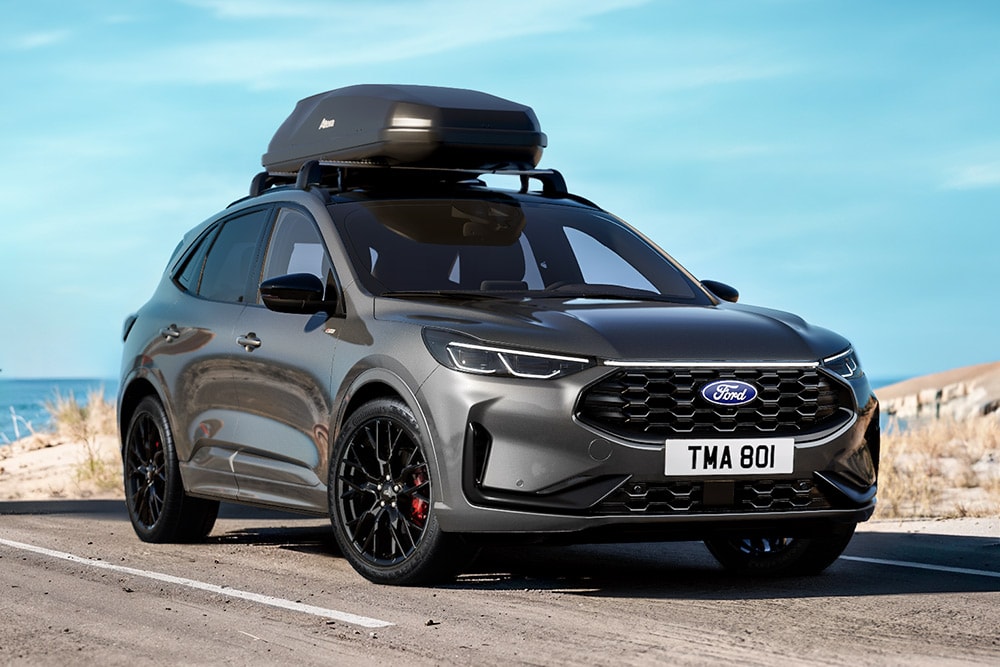 Ford Kuga gris avec coffre de toit, garé sur une route côtière avec l'océan et la plage en arrière-plan.