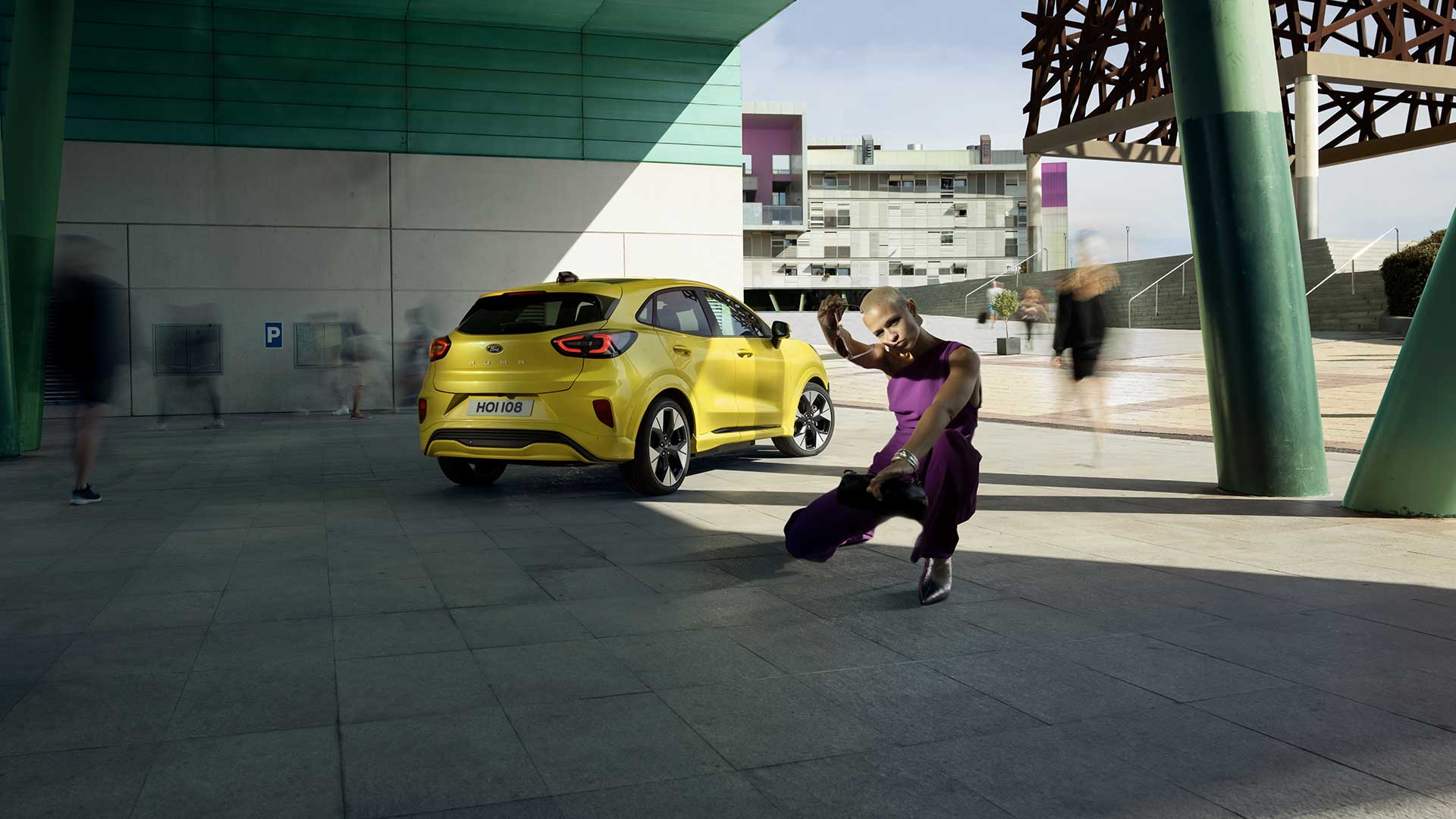 Vue arrière d’un Ford Puma Gen-E® électrique jaune stationné sous un pont, près d’une personne habillée en violet.