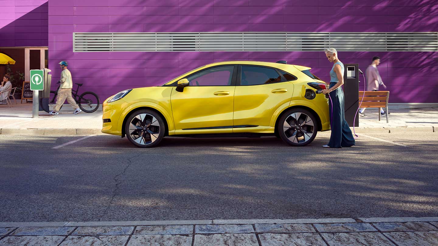 Vue latérale d’un Ford Puma Gen-E® électrique jaune branché à une station de recharge publique sur une place de parking.