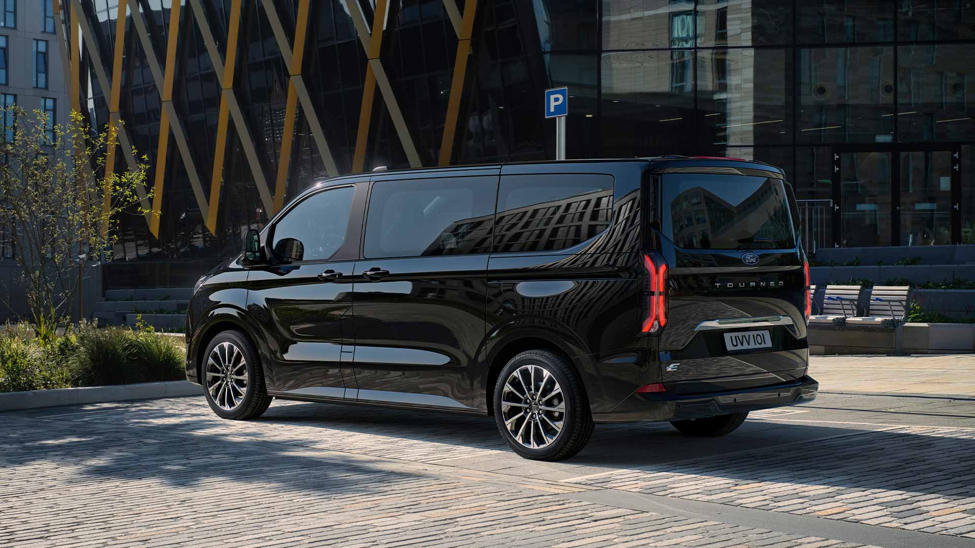 Un Ford E-Tourneo Custom noir est stationné, montrant le côté et l’arrière devant un bâtiment moderne.
