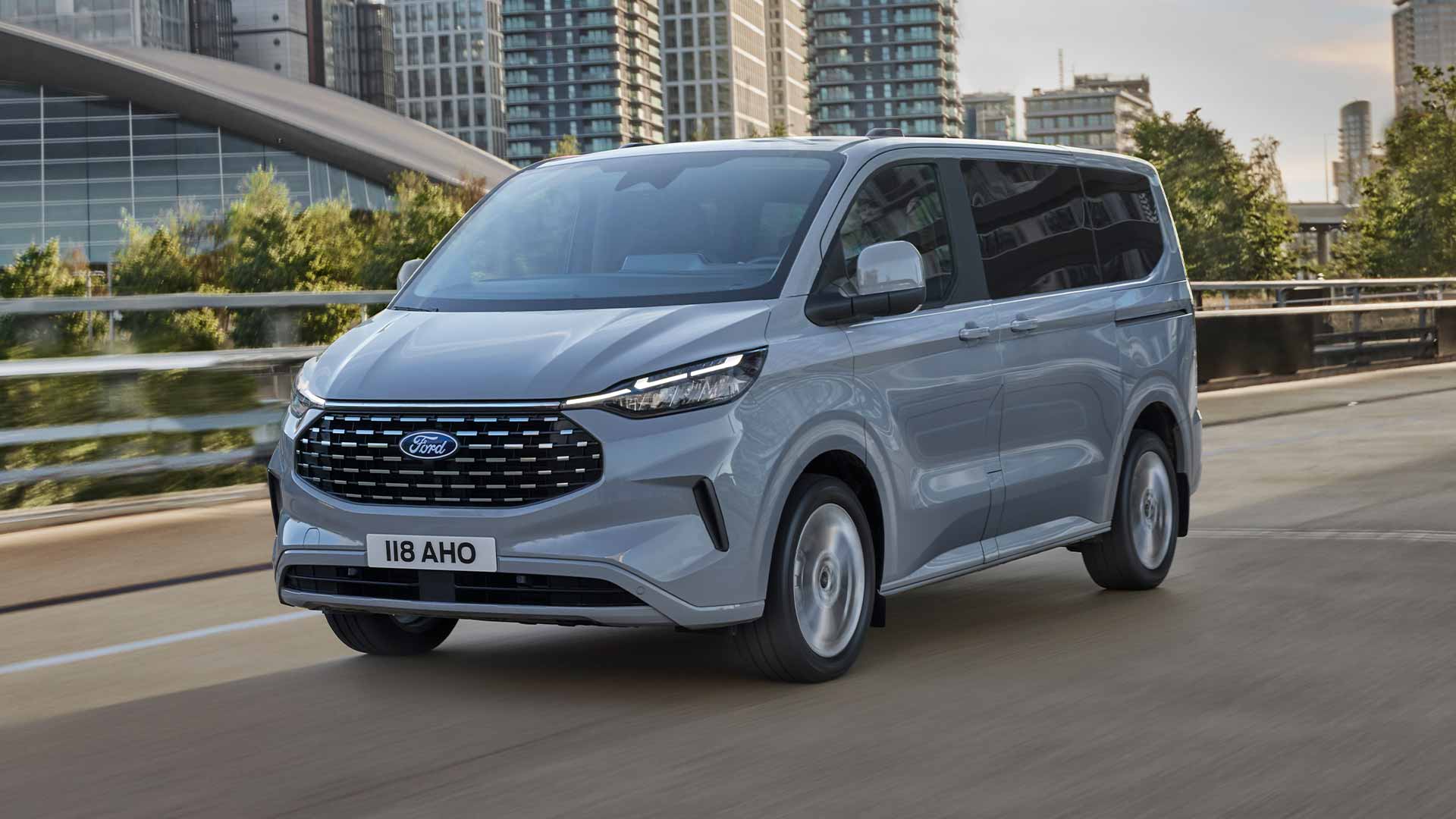 Un Ford Tourneo Custom argenté roule sur une route en ville.