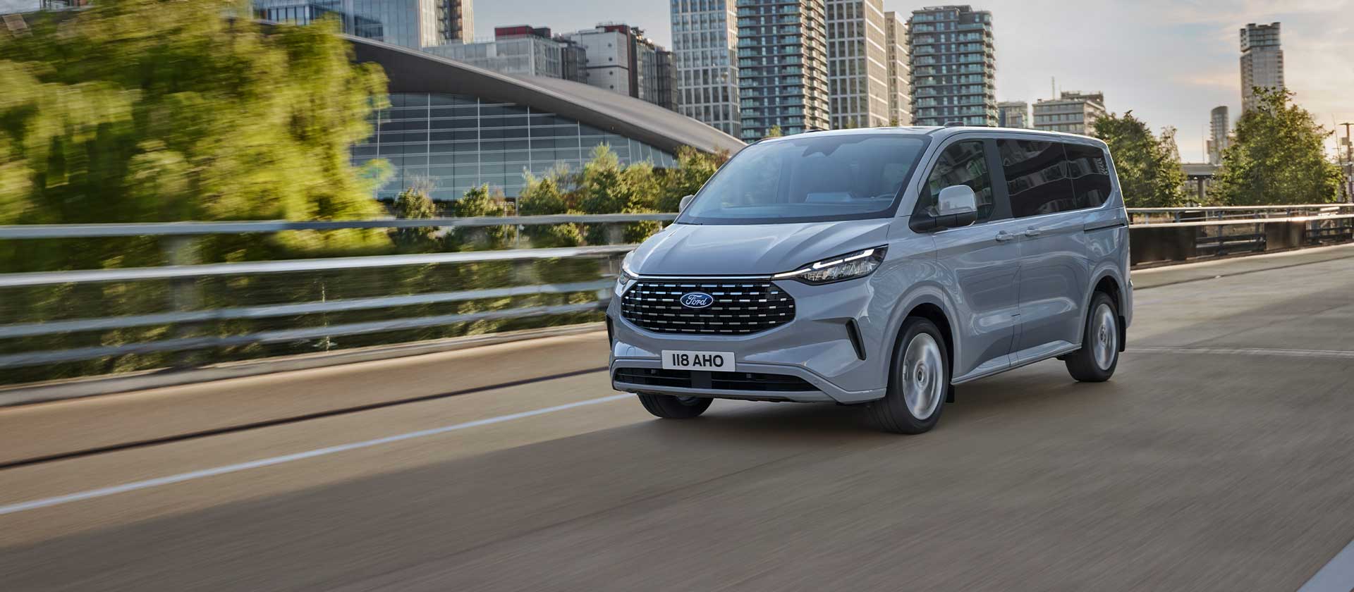 Un Ford Tourneo Custom argenté roule sur une route en ville.