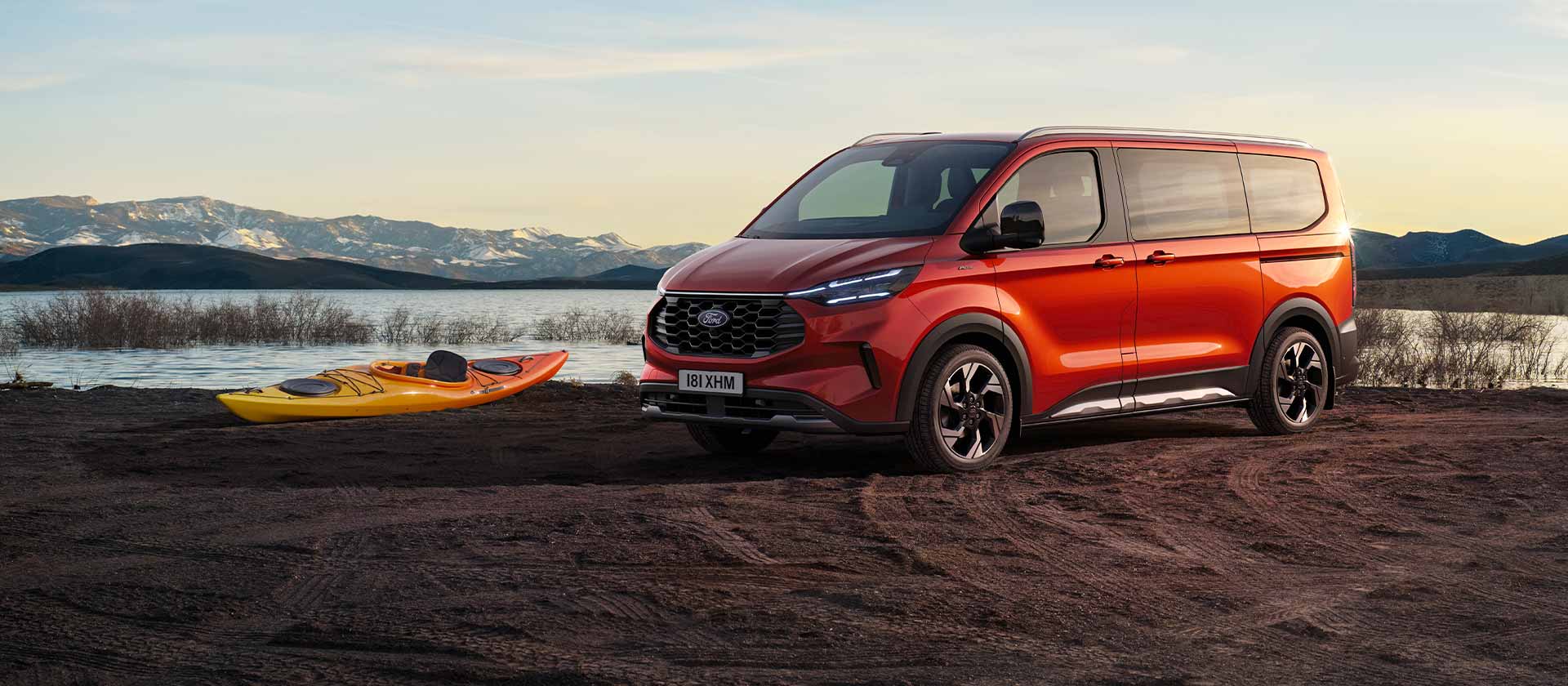 Un fourgon Ford Tourneo Custom rouge est stationné près d'un lac avec un kayak. 
