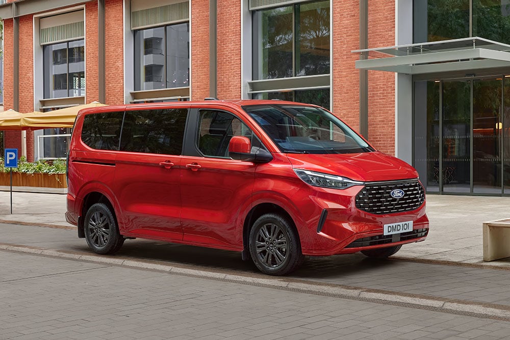 Un Ford Tourneo Custom rouge est stationné dans la rue d'une ville devant un bâtiment en briques