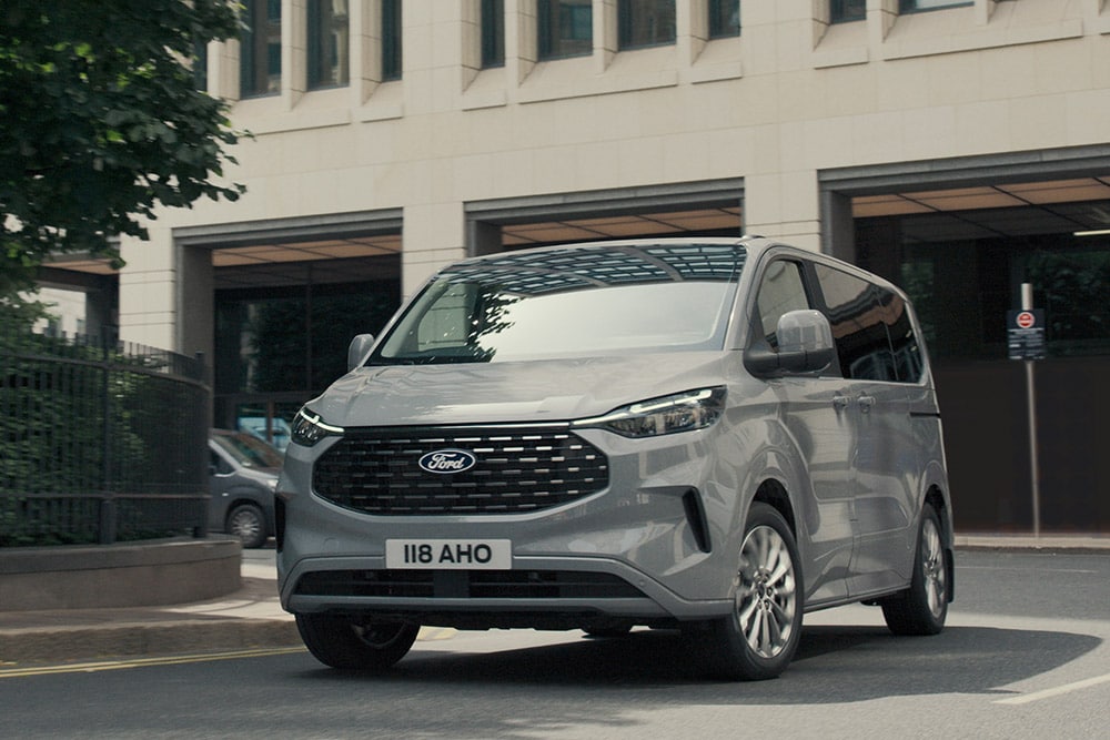 Un Ford Tourneo Custom argenté roule dans la rue d'une ville.