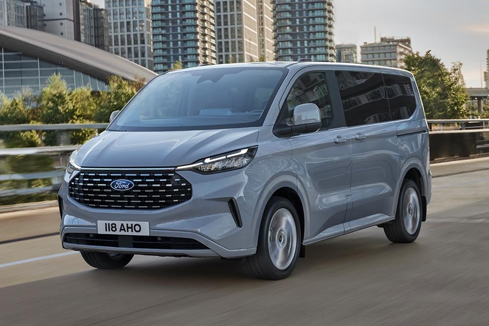 Un Ford Tourneo Custom argenté circule sur une route en ville.