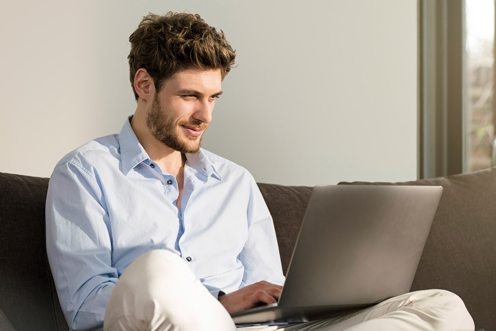 Une personne en chemise bleu clair, assise sur un canapé, utilise un ordinateur portable.