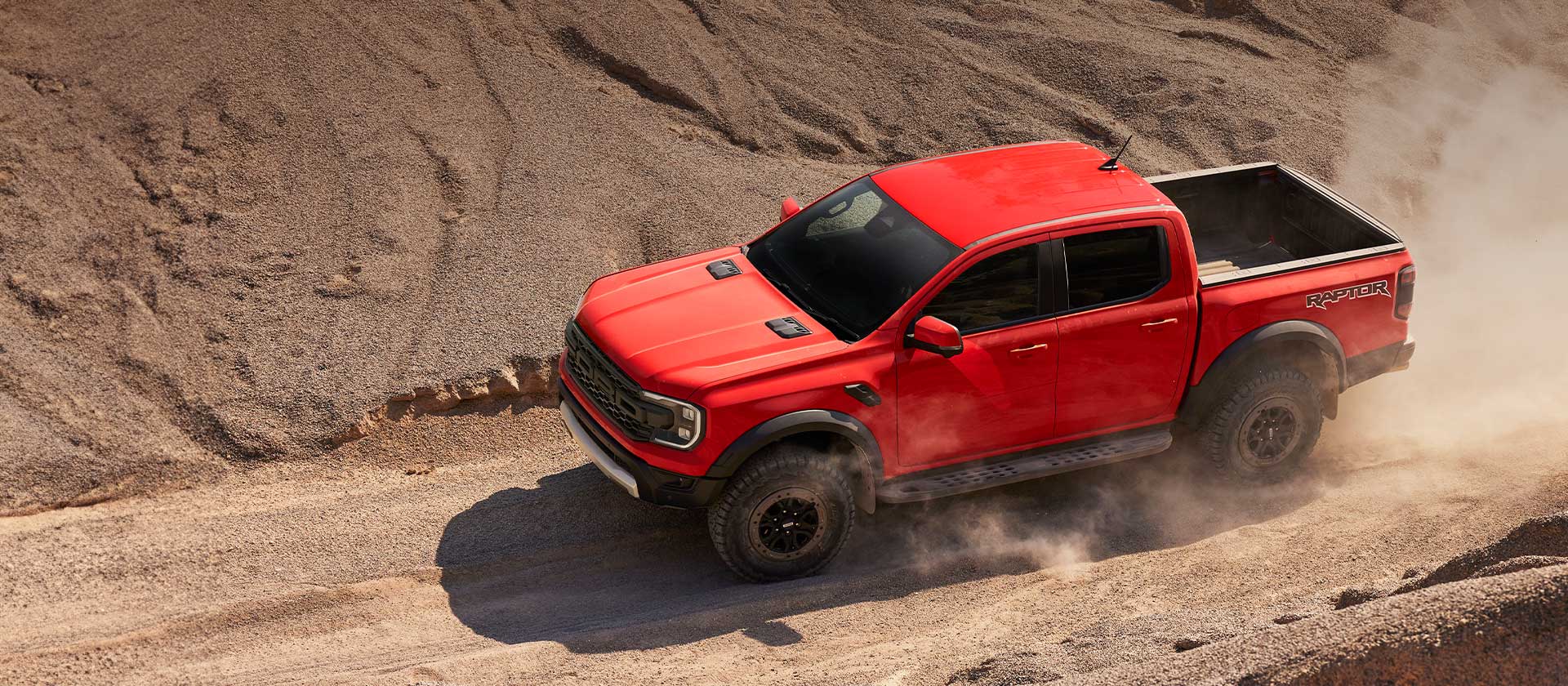 Ford Ranger Raptor orange grimpant une pente dans un nuage de poussière.