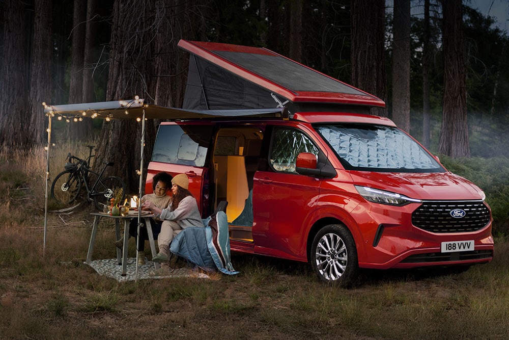 Deux femmes assises sous un auvent fixé aux portes latérales ouvertes d’un Ford Transit Custom Nugget rouge, avec le toit relevable déployé.