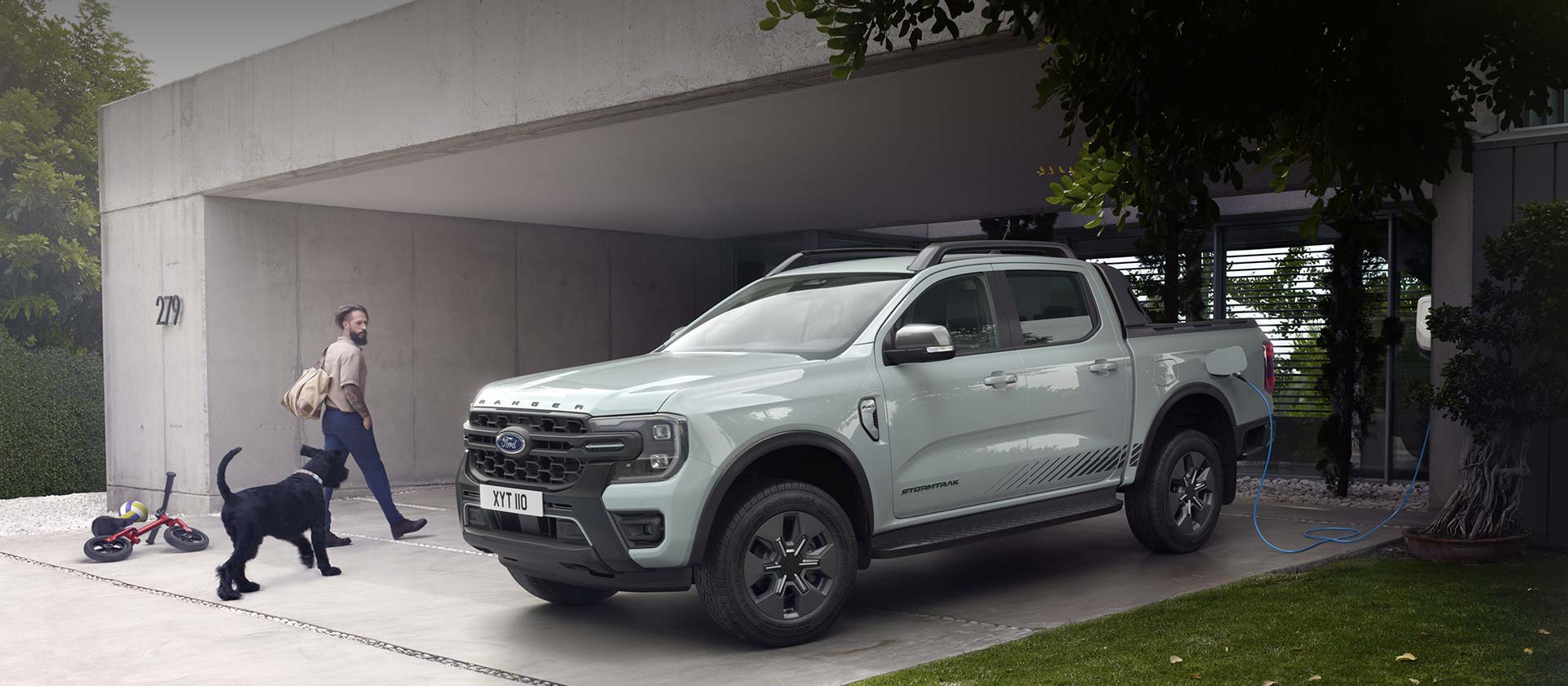 Un homme et son chien entrent dans une maison moderne, passant à côté d'un Ford Ranger Stormtrak Plug-in Hybrid en charge.  (non disponible en Belgique/au Luxembourg)