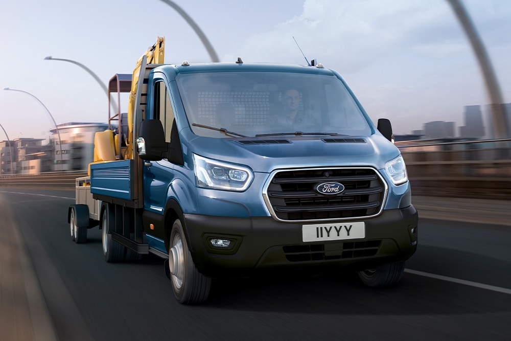 Vue de face de la calandre d’un Ford Transit châssis-cabine transportant une pelleteuse tout en circulant sur une route urbaine. 