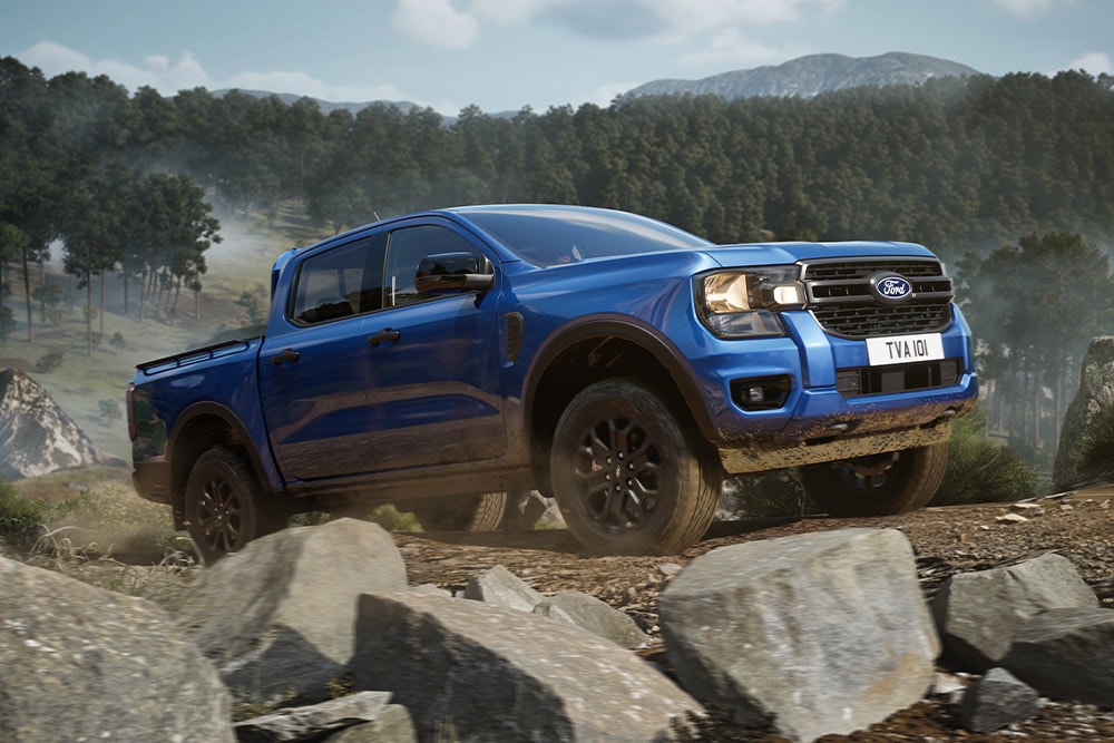 Un pick-up Ford Ranger bleu roule sur un chemin de terre à travers une forêt, laissant une traînée de poussière derrière lui.