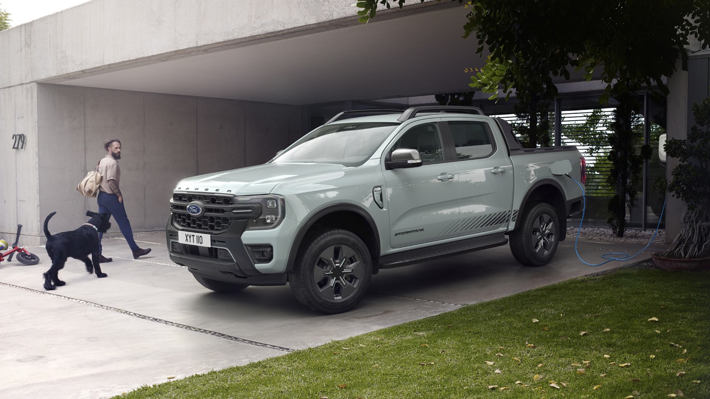 Un homme et son chien entrent dans une maison moderne, en passant devant une Ford Ranger Stormtrak Plug-in Hybrid en charge. 