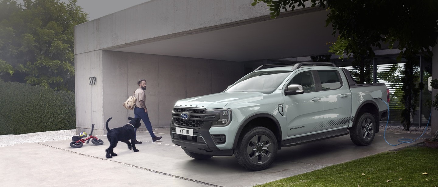 Un homme et son chien entrent dans une maison moderne, en passant devant une Ford Ranger Stormtrak Plug-in Hybrid en charge. 