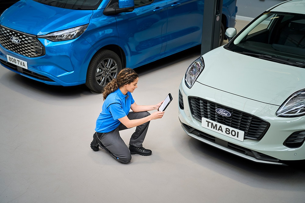 Un technicien agenouillé entre un fourgon Ford bleu et une Ford Puma blanche, regardant une tablette.