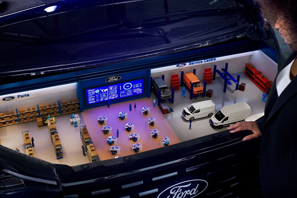 Vue aérienne graphique d'un atelier Ford organisé, avec des étagères de pièces et d'outils en bleu et rouge.