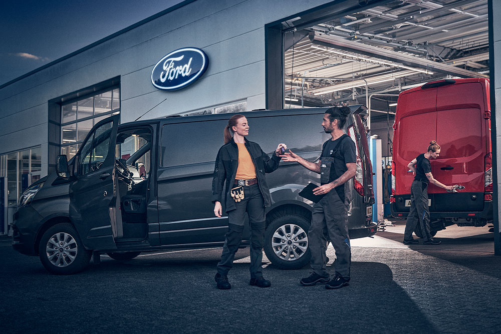 Deux techniciens échangent des clés à l'entrée d'une concession Ford, avec des fourgons garés à l'arrière-plan.