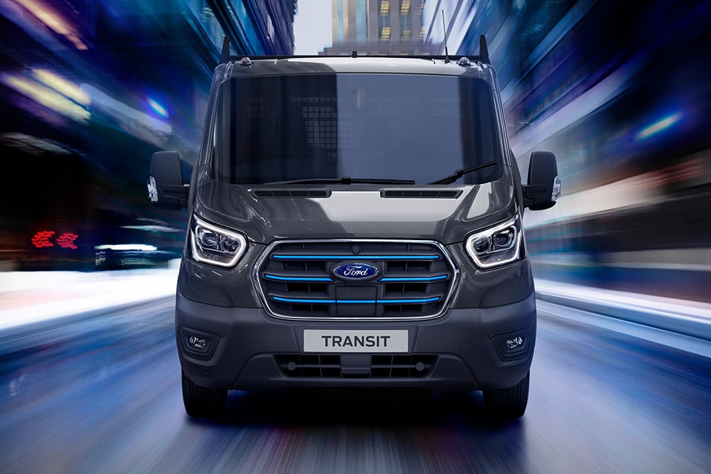 Vue de face d'un fourgon Ford Transit gris foncé en mouvement, avec un arrière-plan flou.