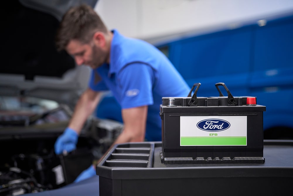 Un mécanicien Ford travaillant sur un moteur de voiture, avec une batterie Ford au premier plan.