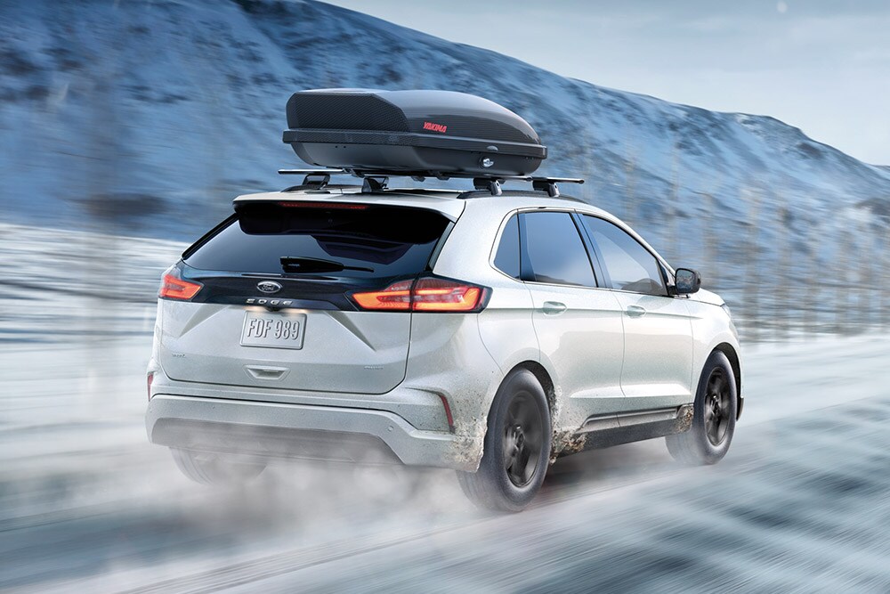  Ford Edge blanc avec coffre de toit roulant sur une route de montagne enneigée, flou de mouvement.
