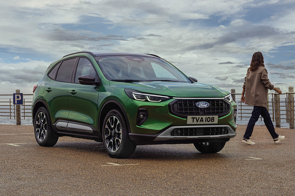 Vue 3/4 avant d'un Ford Kuga vert garé près d'une barrière côtière, avec deux personnes à proximité.